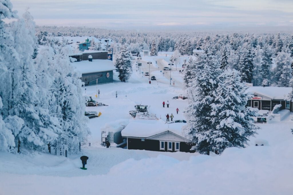 Phần Lan: Thời Tiết Bốn Mùa Xuân - Hạ - Thu - Đông Sẽ Như Thế Nào?! 2 A snowy village surrounded by pine trees covered in snow. Buildings are partially buried, and a few people are visible in the snowy streets. Overcast skies create a serene winter atmosphere.