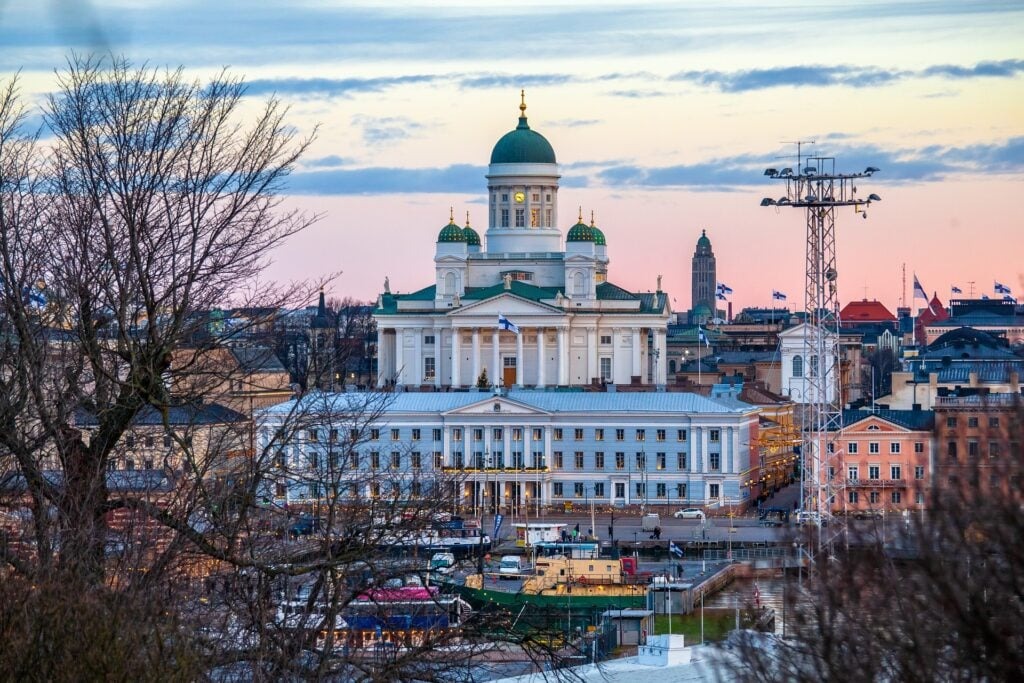 How You Can Earn a Practical Nurse Degree in Finland Without Tuition and Accommodation Fees 4 View of Helsinki Cathedral at sunset with a partly cloudy sky. The scene includes surrounding buildings and a foreground of bare trees, suggesting a winter or early spring setting.