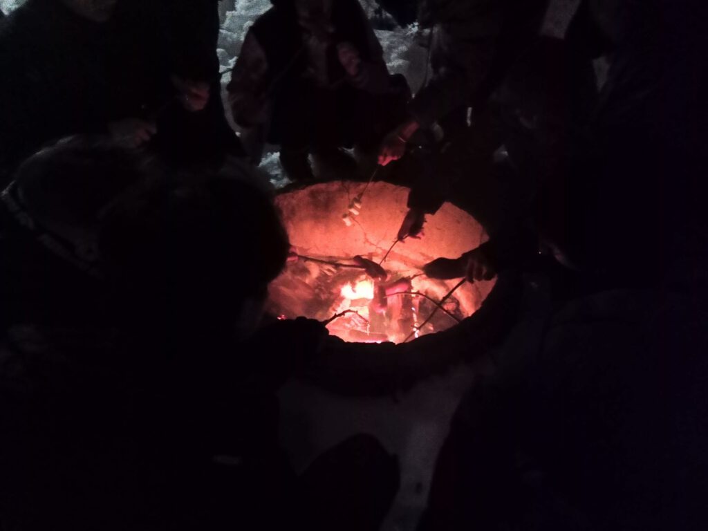 My Firsts in Finland 4 People gather around a fire pit, roasting marshmallows on sticks in the dark. The fire illuminates their hands and the marshmallows, while the surrounding area appears snowy and dimly lit.