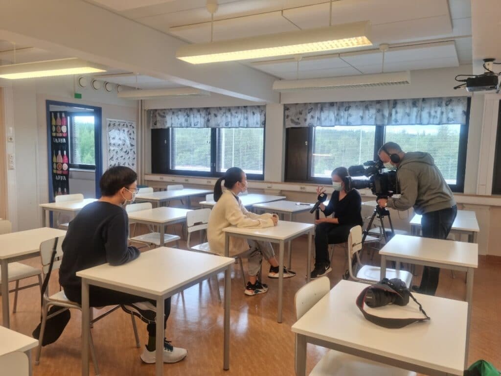 HS2021 Diary: The first Summer Camp ever in Finland! 4 A camera crew films two people sitting at a desk in a brightly lit classroom. The people are wearing masks, and the camera operator is adjusting the equipment. Large windows and empty desks are visible in the background.