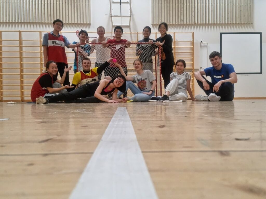 HS2021 Diary: The first Summer Camp ever in Finland! 5 A group of people smiling and posing in a gymnasium, with a few individuals sitting and reclining in the front row. They appear relaxed and cheerful, gathered together in front of a climbing frame and walls lined with wooden panels.