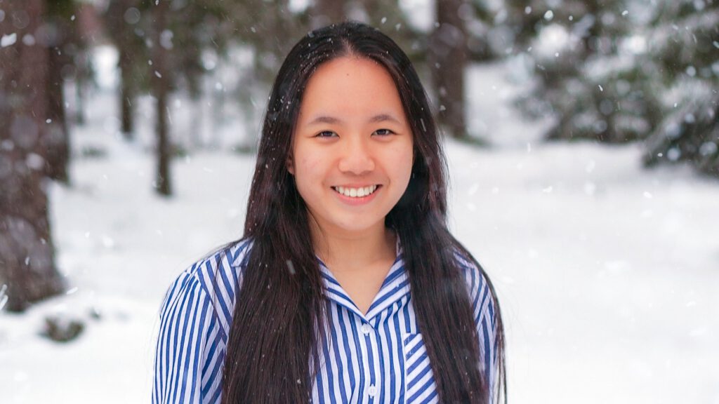 Finding my very own Finest Future in Finland 9 A person with long dark hair, wearing a blue and white striped shirt, stands outdoors in a snowy landscape with trees in the background. Snowflakes are gently falling around them, and they are smiling at the camera.