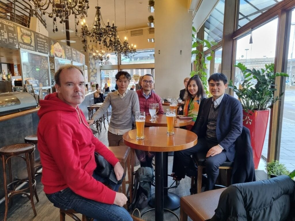 Creating Connectivity - Finest Future's Delegation Trip to Finland 2 A group of five people sitting around a wooden table in a cozy cafe. They are enjoying drinks, including glasses of beer. The cafe has decorative chandeliers and a menu board in the background. Large windows let in natural light from outside.