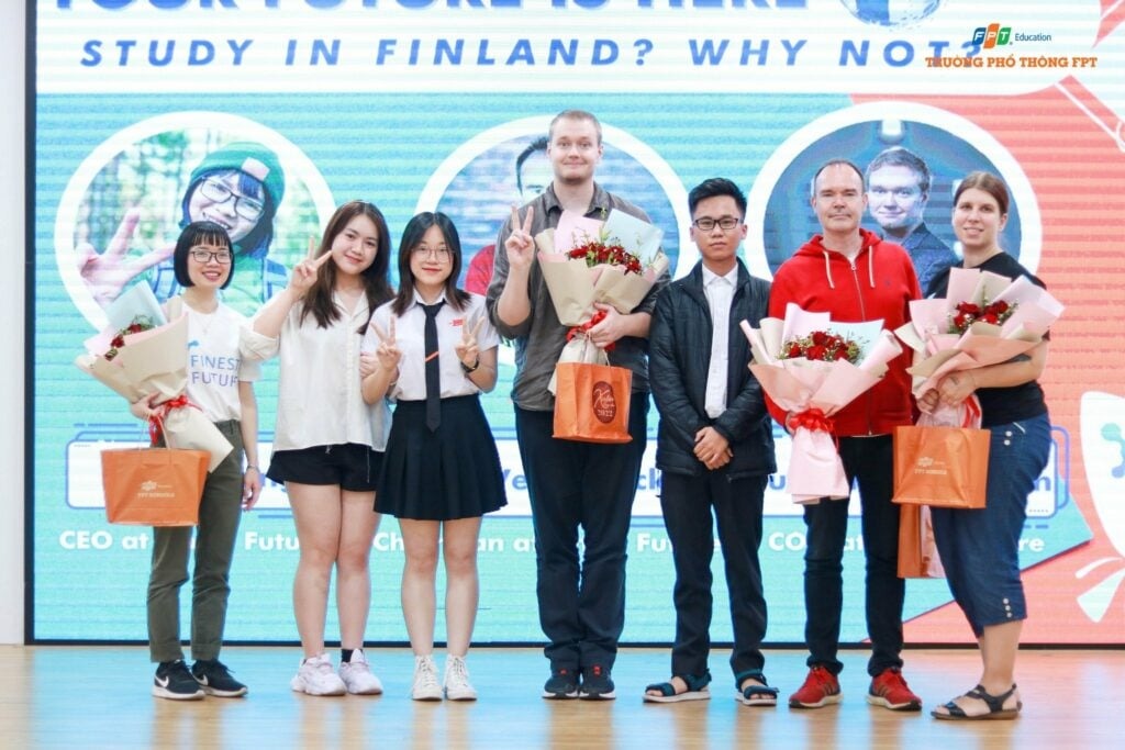 Finest Future’s Delegation Trip to Vietnam - Growth Together, for a Better Future 4 A group of seven people, some holding bouquets and shopping bags, stand smiling on a stage. A background banner reads "YOUR FUTURE IS HERE, STUDY IN FINLAND? WHY NOT?" The atmosphere is positive and celebratory.